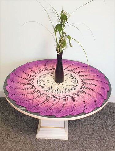 Rundes gehäkeltes Deckchen in Lila, Rosa und Creme mit Spitzenmuster und einer Vase in der Mitte auf einem runden Tisch.