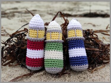 Close-up of three striped crocheted lighthouse toys on sand with seaweed in red, green, and blue.