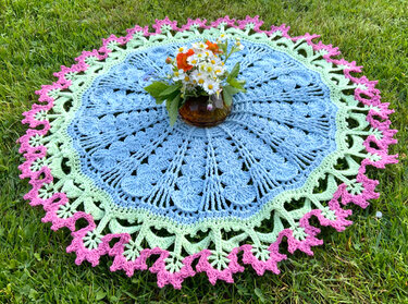 Rundes gehäkeltes Deckchen hellblau mit grüner Bordüre und rosa Spitzenrand, mittig Vase mit Wiesenblumen, liegt auf Gras