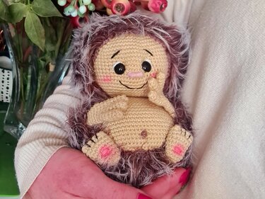 Gehäkelter Amigurumi-Igel mit beigem Körper, braunem flauschigem Fell und rosa Wangen, gehalten in zwei Händen.