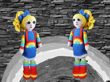 Zwei gehäkelte Amigurumi-Puppen mit blonden Zöpfen, blauem Kleid, Regenbogen-Details und roten Stiefeln vor Steinwand