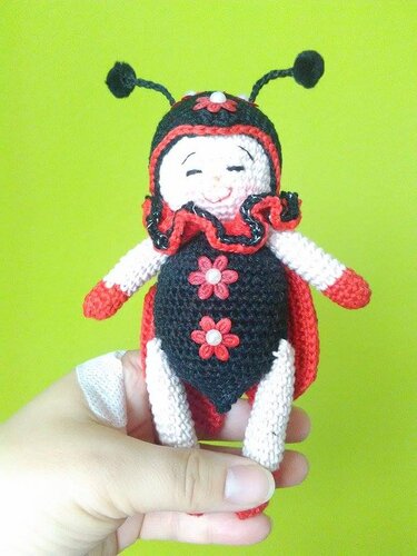 Gehäkelter Marienkäfer-Glücksbringer mit Rüschenkragen, Blumenverzierung und Antennen, gehalten von einer Hand