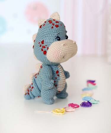 Gehäkelter hellblauer Amigurumi-Dinosaurier mit beiger Schnauze und roten Punkten neben bunten Häkelherzen