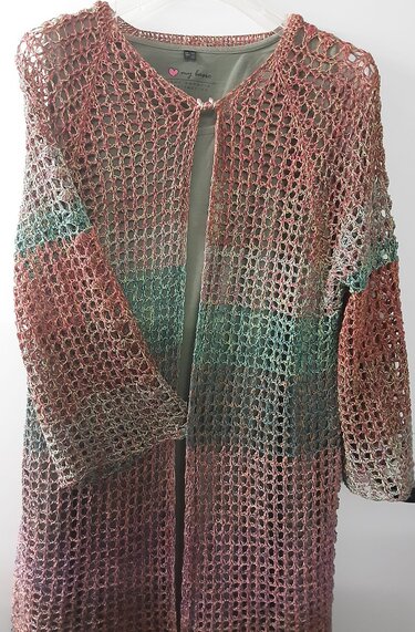 Lange offene Häkeljacke in Rosé, Grün und Violett mit lockerem Netzmuster, über einem olivgrünen Shirt an einem Kleiderbügel hängend.