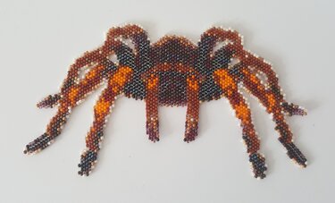 Spinnenmotiv aus kleinen braunen, orangefarbenen und schwarzen Perlen auf weißem Hintergrund.