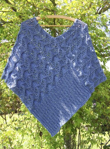 Blauer gestrickter Damen-Poncho mit V-Ausschnitt und Ajourmuster an einem Bügel vor grünen Sträuchern