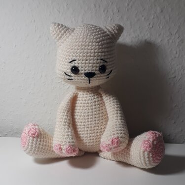 Cremefarbene Amigurumi-Katze mit rosa Pfotenballen, schwarzen Augen und gestickten Schnurrhaaren, sitzend.