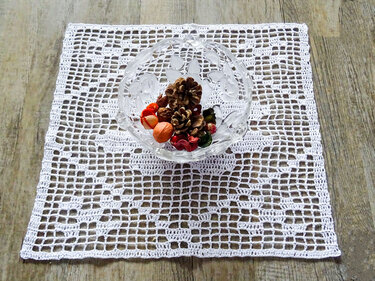 Quadratisches weißes gehäkeltes Filetdeckchen auf Holztisch mit Glasschale und getrockneten Deko-Elementen