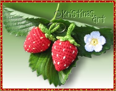 Nahaufnahme zweier gehäkelter Erdbeeren mit weißen Stichen, grünen Kappen, Metallringen und einer kleinen weißen Häkelblüte