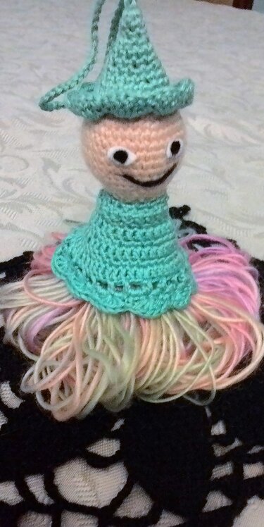 Amigurumi‑Püppchen mit mintgrünem Hut und Rock aus pastellfarbenen Garnfransen auf dunklem Häkeluntergrund