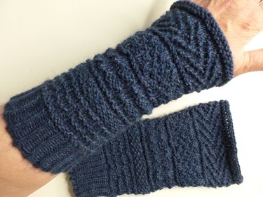 Dunkelblaue, gestrickte Armstulpen mit diagonalem Rippenmuster, eine am Unterarm getragen, die zweite daneben liegend