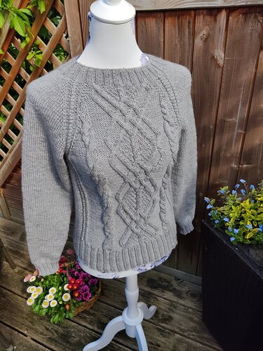 Gestrickter grauer Pullover für Damen mit Zopfmuster auf einer Schneiderpuppe vor Holzzaun