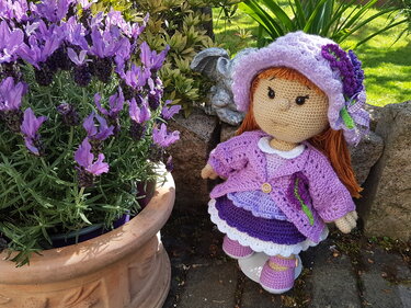 Gehäkelte Amigurumi-Puppe mit lila Kleid und Blumenhut neben blühendem Topf