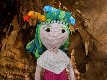 Gehäkelte Amigurumi‑Medusa‑Puppe mit bunten Schlangenhaaren und weißem Schal vor Höhlenwand