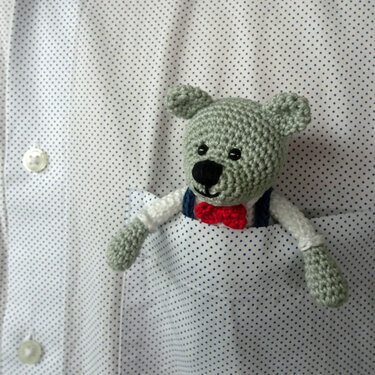 Gehäkelter graugrüner Amigurumi-Teddy mit roter Fliege in gepunkteter Hemdtasche