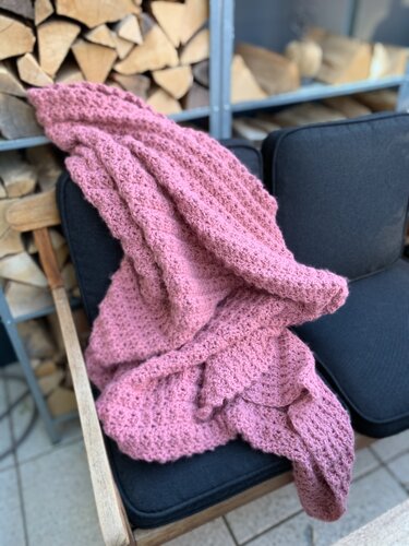 Rosafarbene Häkeldecke mit ausgeprägtem Muster auf dunklem Stuhlkissen vor Regal mit Brennholz.