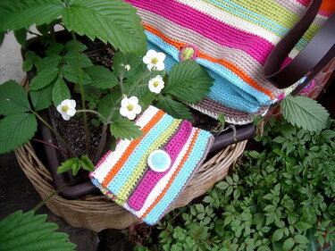Gehäkelte gestreifte Tasche und passende Geldbörse im Weidenkorb neben Erdbeerpflanzen mit weißen Blüten