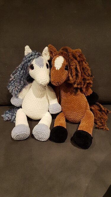 Zwei gehäkelte Amigurumi-Pferde, weiß mit blau-grauer Mähne und hellblauen Hufen, braun mit kastanienbrauner Mähne und schwarzen Hufen, auf dunklem Hintergrund.