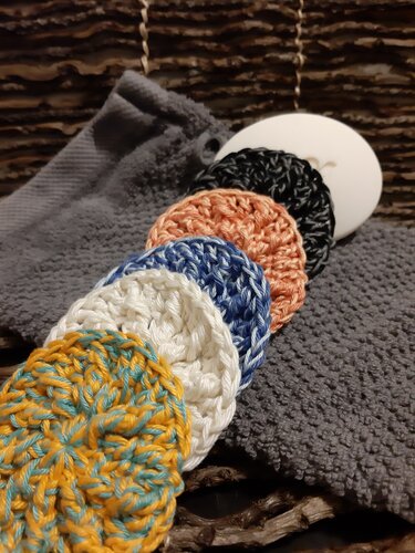 Fünf runde, gehäkelte Pads in Gelb, Weiß, Blau, Pfirsich und Schwarz auf einem grauen Frottiertuch.