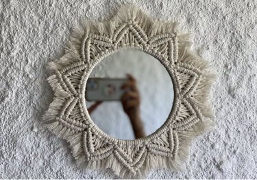 Runder Wandspiegel mit naturfarbenem Makramé‑Rahmen in Sonnensternform an weißer Wand; Hand mit Smartphone spiegelt sich im Glas.
