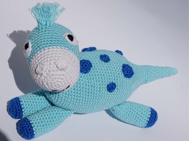 Light blue crocheted amigurumi dinosaur with dark blue spots, dark blue toe tips, a white snout, black eyes, and a tuft of yarn on its head, on a white background.