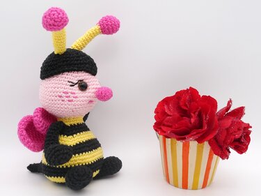 Gehäkelte Amigurumi-Biene in Schwarz-Gelb mit rosa Kopf und Flügeln neben roten Rosen im gestreiften Becher
