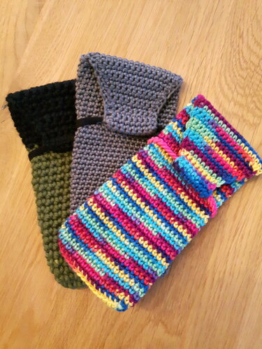 Drei handgehäkelte Hüllen in Grau, Olivgrün und bunten Streifen auf Holz