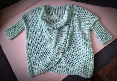 Mintgrüner gehäkelter Damen-Cardigan mit Schalkragen und 3/4-Ärmeln liegend auf rosa Untergrund