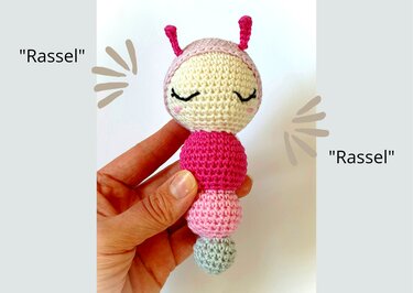 Hand hält gehäkelte Babyrassel in Pink, Hellrosa, Grau und Creme.