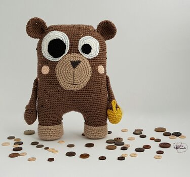 Brauner gehäkelter Amigurumi-Teddybär mit großen Augen, beigen Details und gelber Tasche, umgeben von Knöpfen