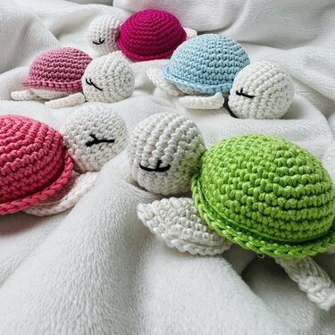 Mehrere gehäkelte Schildkröten-Rasseln in verschiedenen Farben auf weißer Decke