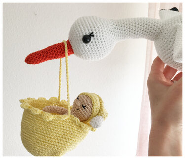 Gehäkelter weißer Storch mit orangefarbenem Schnabel hält gelbes Körbchen mit gehäkeltem Baby