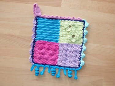 Gehäkelte Baby-Fühldecke aus vier pastellfarbenen Quadraten mit unterschiedlichen Strukturen, Aufhängeschlaufe und Fransen.