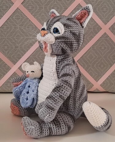 Sitzende grau-weiße Amigurumi-Katze mit blauen Augen und kleiner weißer Maus im blauen Tuch.