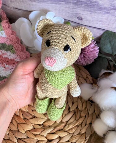 Gehäkelter Amigurumi-Teddybär in Beige mit rosa Nase, grünem Halstuch und grünen Schuhen, der in einer Hand gehalten wird