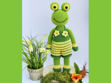 Crocheted green frog in yellow-striped romper with flower appliqués beside potted plant