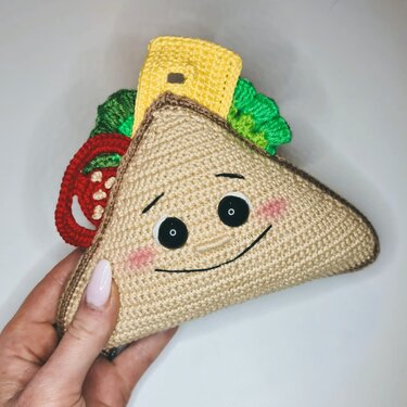 Hand hält gehäkeltes dreieckiges Sandwich-Amigurumi mit schwarzen Knopfaugen, rosa Wangen und gehäkelten Tomaten-, Salat- und Käse-Applikationen