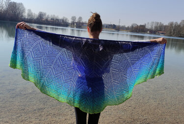 Person hält ausgestrecktes halbrundes Tuch mit blau‑grünem Farbverlauf vor einem See