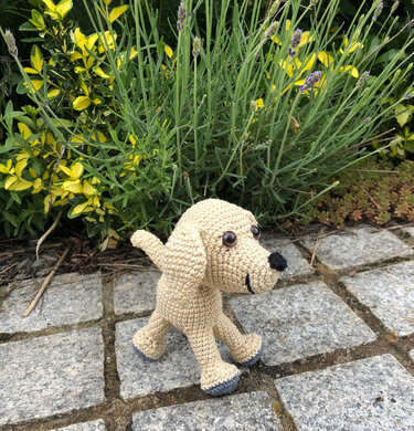 Beiger gehäkelter Amigurumi-Hund mit grauen Pfoten auf Steinpflaster vor Büschen