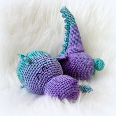 Gehäkelter Amigurumi-Drache in lila und türkis auf weißer flauschiger Decke