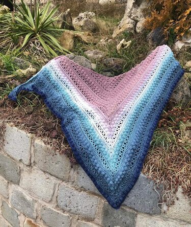 Gehäkeltes Dreieckstuch mit Farbverlauf in blau, türkis, weiß und rosa, auf einer Steinmauer und Gras drapiert.