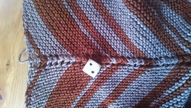 Gestricktes Dreieckstuch mit diagonalen Streifen in Braunrot und Graublau auf einer Holzoberfläche, mit einem kleinen weißen Würfel darauf.