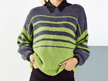 Oversize-Pullover mit breiten lindgrünen und dunkelgrauen Streifen; schwarzer Hemdkragen sichtbar, Hände in den Taschen.
