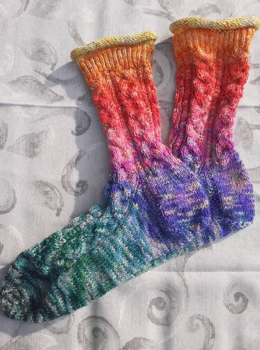 Paar handgestrickter Socken mit Farbverlauf von Grün über Violett zu Orange und Gelb, mit Zopfmuster und geripptem Bündchen, liegend auf hellem Stoff