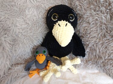 Large black crocheted crow with cream beak and glossy eyes next to a small grey crocheted bird with an orange beak, on a fluffy grey blanket.