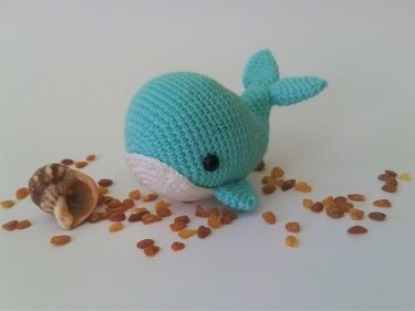 Compact turquoise crochet whale with white underside and visible stitch texture, photographed with a shell and amber beads on a white background