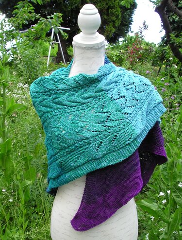 Gestricktes Dreieckstuch in Türkis bis Violett mit Zopf- und Lochmustern auf einer Schneiderpuppe im Garten.