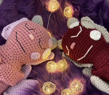 Zwei gehäkelte Amigurumi‑Drachen (rosa und bordeaux) auf lila Kunstfell mit Lichterkette