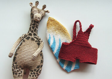 Beige and brown crocheted giraffe amigurumi with yellow-white-blue striped surfboard and small red swimsuit on a light background.