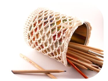 Umhäkelte Dose als Utensilo, liegend und gefüllt mit Buntstiften und Pinseln; offenes Häkelnetz mit beigefarbenem Band.